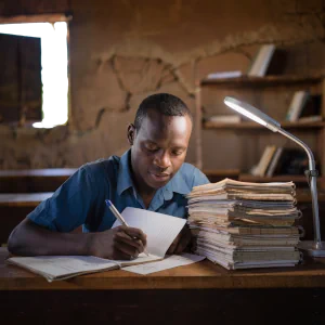 Teacher reviewing handwritten student notebooks in a poorly stocked African school library. Illustrates the challenge of providing individual feedback and personalised learning in resource-constrained settings without digital support.